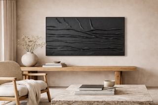 A warm, minimalist living space with a large black textured abstract artwork mounted above a wooden console table, complemented by neutral furnishings, ceramic decor, and soft natural light.