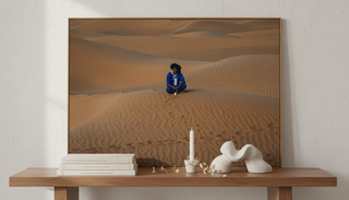 A framed landscape photograph depicting a person seated on rippled desert sand dunes, styled in a warm minimalist interior with a wooden console, stacked books, a candle, and abstract sculptural decor.