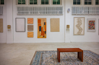 A contemporary art gallery featuring a series of abstract and textured artworks in warm earth tones arranged along a white wall, with a wooden bench centered on a patterned rug in the foreground.